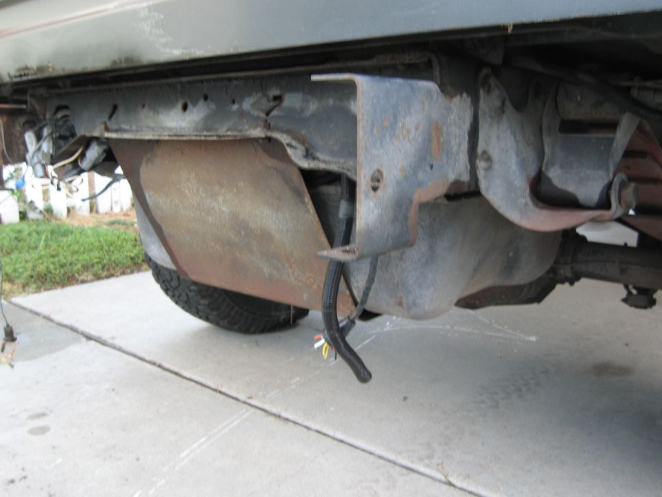 Underside view of a vehicle's frame and gas tank, with some wiring visible. A tire is partially seen