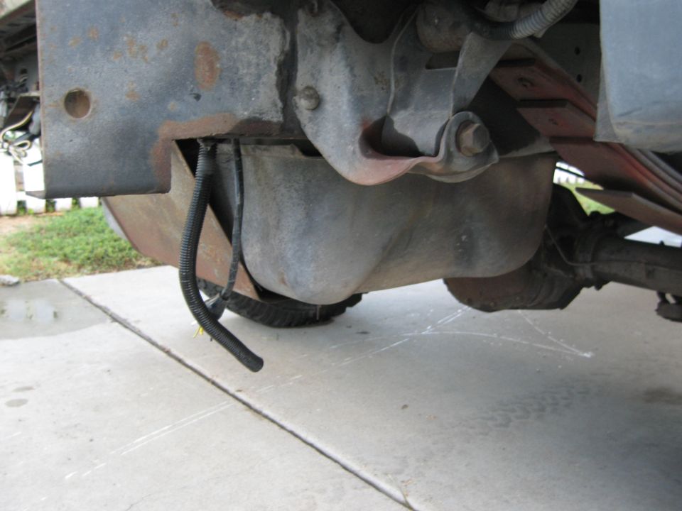 Underside view of a vehicle's gas tank and leaf spring suspension, with black corrugated tubing hanging