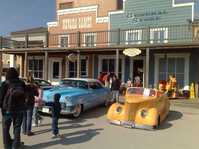 Yellow convertible hot rod and blue two-door sedan parked outside a general store