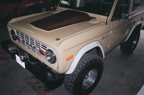 1976 Ford Bronco - Front 3/4 view of a tan 1976 Ford Bronco, modified with a hood scoop and off-road tires.