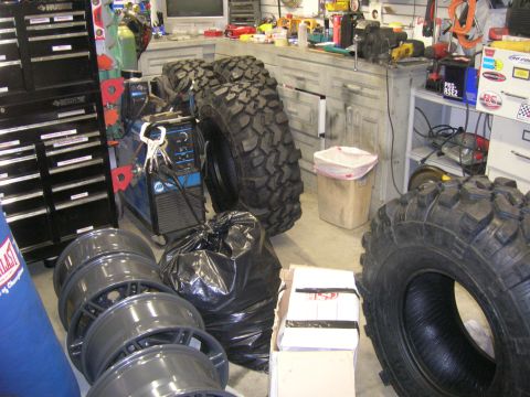 1996 Ford Bronco - A cluttered garage scene displays large off-road tires, a stack of rims, and welding equipment, suggesting preparation for vehicle modifica…