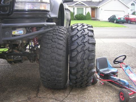 1996 Ford Bronco - Two mud-terrain tires stood upright side by side on a driveway for a tread and size comparison, the taller aggressive-lug tire on the right…