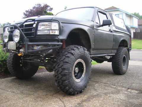 1996 Ford Bronco - Dark gray Ford Bronco built for off-road, shown from a low three-quarter front angle on a driveway. It has a custom tube front bumper with…