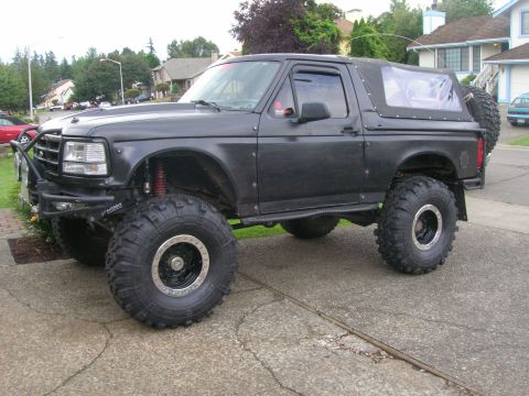 1996 Ford Bronco - A matte black 1990s Ford Bronco two-door SUV with a soft top, shown from a 3/4 front angle. It has a large suspension lift, oversized off-r…