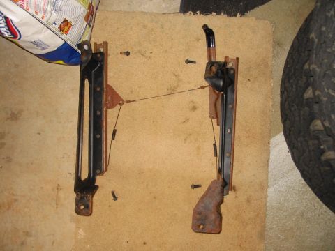 1988 Ford Bronco - A pair of removed seat slide rails laid out on cardboard on a garage floor, still joined by the release linkage rod and cables, with a blac…