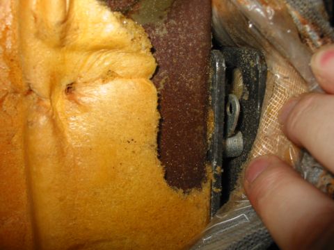 1988 Ford Bronco - Extreme close-up of a seat-back attachment bracket with fingers holding the burlap backing aside, showing the rusted frame tube, the slotte…