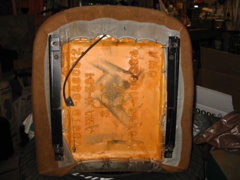 1988 Ford Bronco - A brown cloth bucket seat cushion flipped upside down on a tire in a workshop, with the burlap backing exposed and orange foam stamped 'AWA…