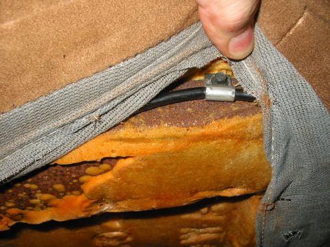 1988 Ford Bronco - Close-up of a seat teardown with a hand holding back the gray scrim and tan upholstery to reveal orange foam over a rust-streaked stamped-s…
