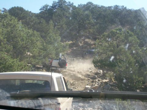 1993 Ford Bronco - Multiple SUVs tackle a challenging, dusty, rocky trail in a wooded area.