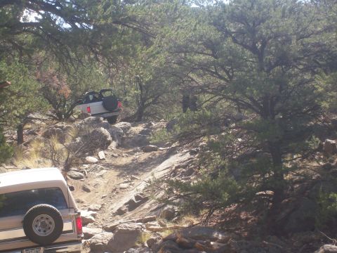 1993 Ford Bronco - Two SUVs tackle a challenging off-road trail in a wooded, rocky area.