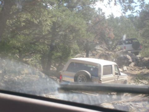 1993 Ford Bronco - Two Ford Broncos navigate a challenging off-road path through a wooded, rocky terrain.