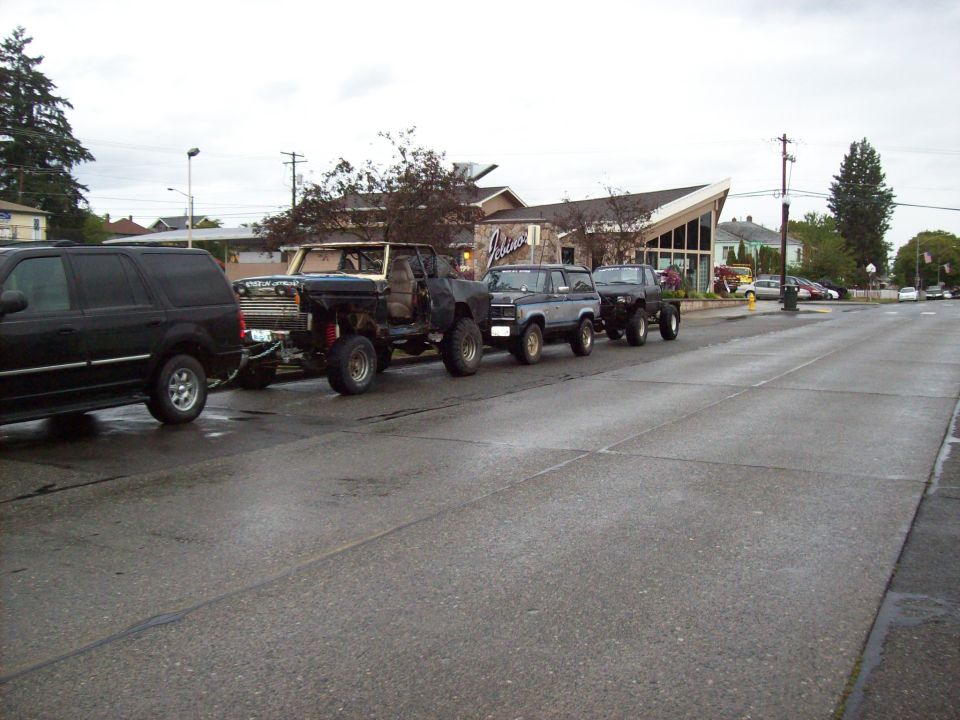 Black SUV towing a heavily modified truck, followed by two more trucks on trailers