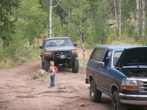 1993 Ford Bronco - Two Ford Broncos, one brown and lifted, the other blue with hood open, sit on a dirt path surrounded by trees.