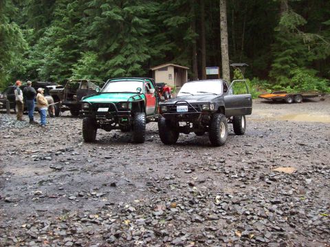 1985 Toyota Pickup/SR5 - Two heavily modified Toyota Pickup SR5 trucks are parked off-road in a wooded area.