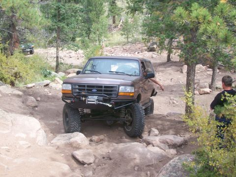 1993 Ford Bronco - A brown Ford Bronco is shown off-roading on a rocky trail with another vehicle visible in the background.