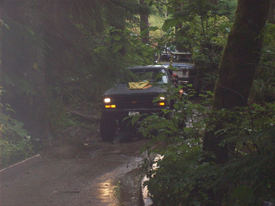 Several trucks driving on a muddy, wooded trail. One truck in the foreground has its headlights on