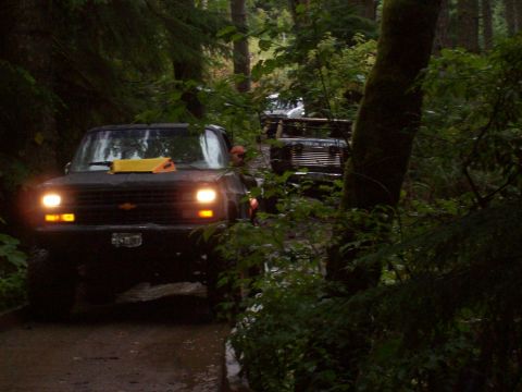 1985 Toyota Pickup/SR5 - A line of off-road vehicles navigates a wooded trail, with the lead truck's headlights cutting through the dim light.