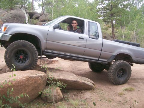 1993 Ford Bronco - A silver pickup truck is shown off-roading over large rocks on a dirt trail.
