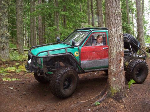 1985 Toyota Pickup/SR5 - A heavily modified pickup truck with a roll cage and oversized tires is seen off-road in a forest setting.