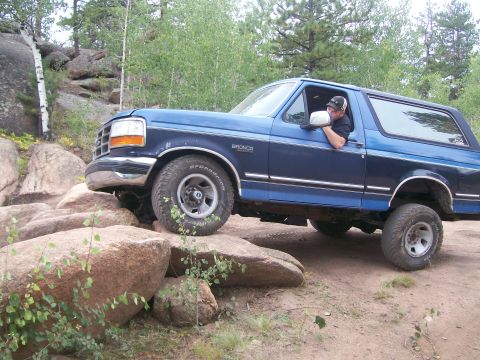 1993 Ford Bronco - A blue 1993 Ford Bronco is shown off-roading over large rocks on a dirt path.