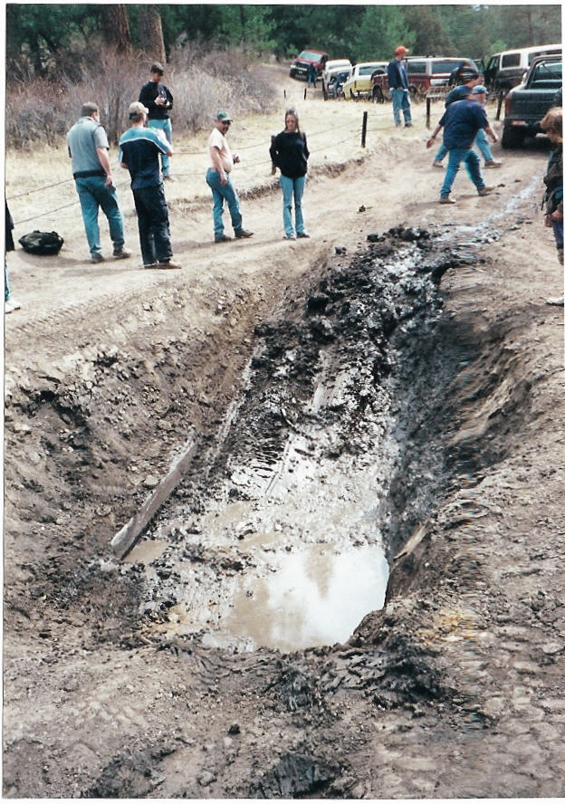 Group of people standing around a large muddy water-filled trench on a dirt trail with parked rigs
