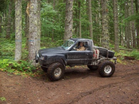 1985 Toyota Pickup/SR5 - A heavily modified pickup truck navigates a wooded trail, its off-road capabilities.