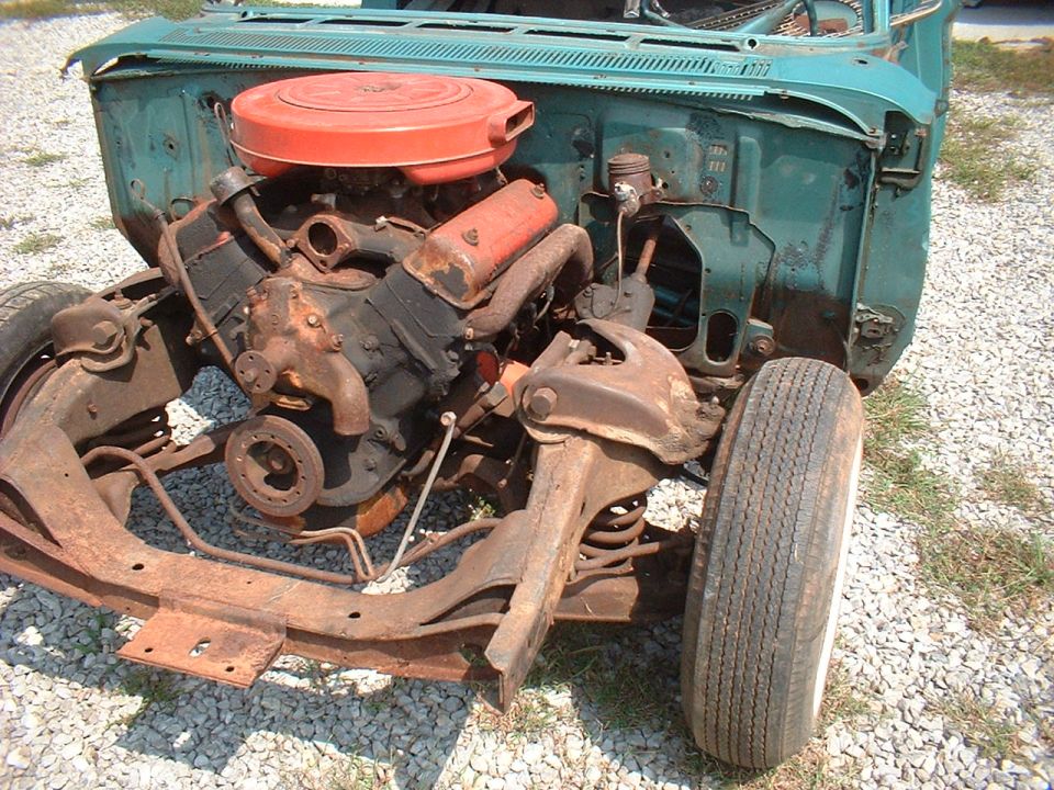Teal vehicle front end stripped down, V8 with red air cleaner, exposed suspension on gravel
