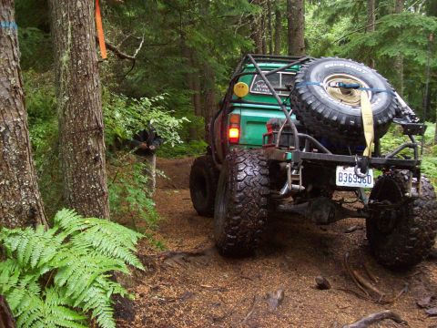 1985 Toyota Pickup/SR5 - A modified green pickup truck navigates a wooded trail, its off-road capabilities with large tires and a rear-mounted spare.