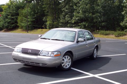 2001 Ford Crown Victoria - A silver 2004 Mercury Grand Marquis is parked in an empty parking lot with trees in the background.