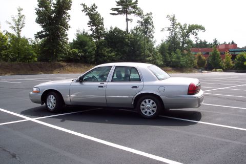 2001 Ford Crown Victoria - Silver sedan parked in an empty lot, side view, with trees and a building in the background.