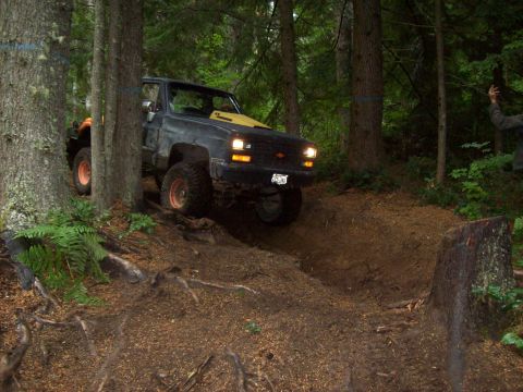 1985 Toyota Pickup/SR5 - A modified Chevrolet pickup truck navigates a challenging off-road course amidst dense woodland.