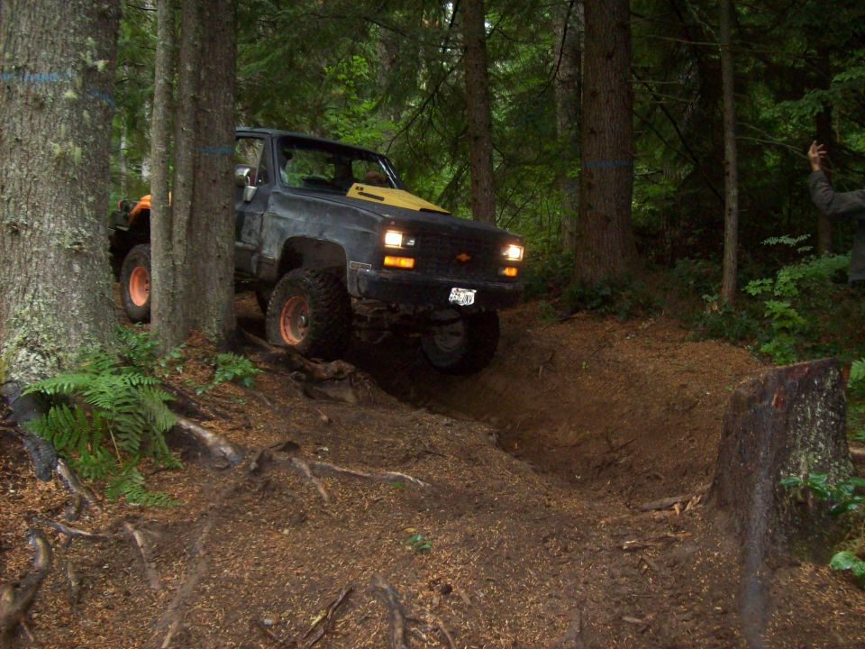 Dark Chevrolet pickup truck with yellow hood decal, orange wheels, and off-road tires, climbing a dirt hill in a forest