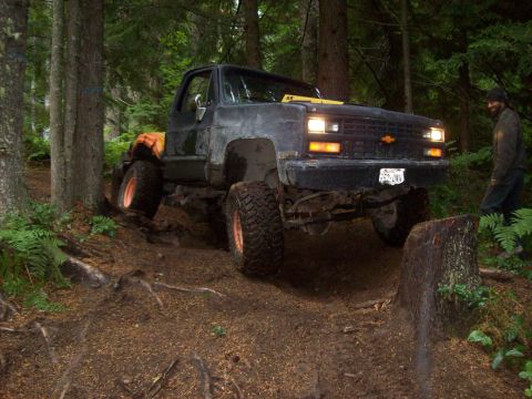 1985 Toyota Pickup/SR5 - A lifted black pickup truck is shown off-roading on a dirt path surrounded by trees.