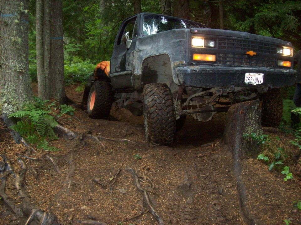 Black Chevrolet pickup truck with large tires driving on a dirt trail in a forest. License plate reads 752-UNV