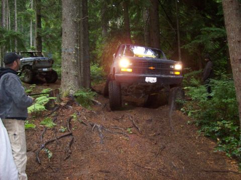 1985 Toyota Pickup/SR5 - Two trucks navigate a muddy, root-covered trail through a dense forest with sunlight filtering through the trees.