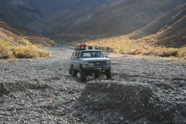 Gray Chevrolet Suburban with roof rack driving on a rocky, dry creek bed