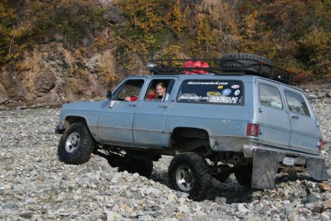 1990 Chevrolet Suburban - A lifted 1990 Chevrolet Suburban navigates a rocky terrain, equipped for off-road adventure with extra fuel and a spare tire.