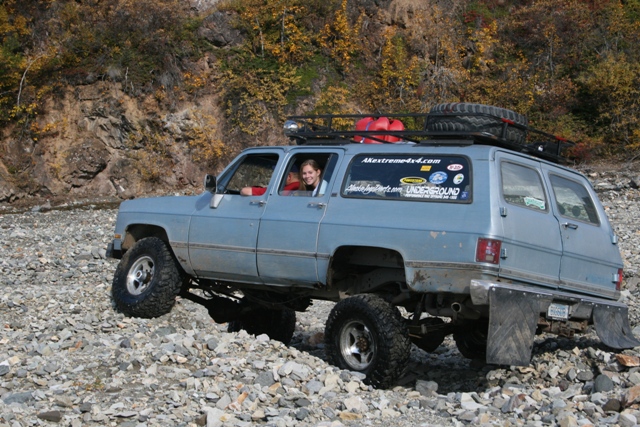 Light blue lifted 1990 Chevrolet Suburban driving on a rocky trail with a woman in the driver's seat. It has a roof rack with gas cans and a spare tire