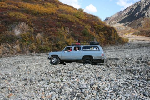 1990 Chevrolet Suburban - A 1990 Chevrolet Suburban navigates a rocky, dry riverbed during autumn, with colorful foliage on the surrounding hills.