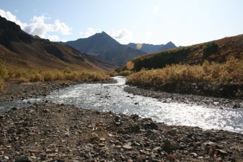1990 Chevrolet Suburban - A wide, sunlit river flows through a rocky valley with mountains in the background.