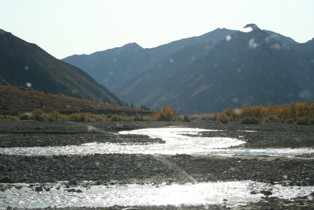 Rocky riverbed with water flowing through it, backed by hills and mountains under a hazy sky