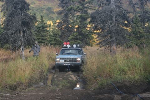 1990 Chevrolet Suburban - A person stands in tall grass next to an off-road vehicle navigating a muddy trail.
