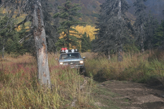 Silver Chevrolet Suburban with roof rack and lights driving on a dirt path through tall grass and trees