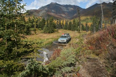 1990 Chevrolet Suburban - A light blue SUV navigates a muddy stream in a mountainous, wooded area during autumn.