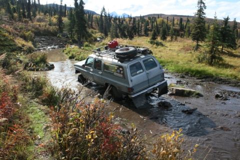 1990 Chevrolet Suburban - An off-road vehicle navigates a challenging muddy terrain with trees and hills in the background.