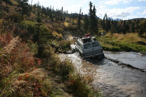 1990 Chevrolet Suburban - A gray van navigates a muddy stream in a wooded, autumnal landscape.