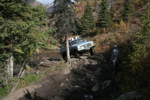 1990 Chevrolet Suburban - SUV tackling a challenging muddy and rocky off-road trail surrounded by trees.