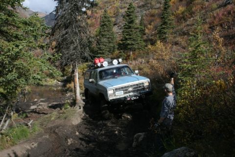 1990 Chevrolet Suburban - An off-road vehicle tackles a challenging, muddy path surrounded by autumn foliage and trees.