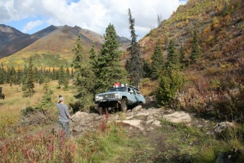 1990 Chevrolet Suburban - A person photographs a light blue SUV as it traverses a challenging rocky terrain in a mountainous, wooded area.