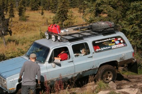 1990 Chevrolet Suburban - This light blue SUV, equipped with a roof rack, spare tire, and auxiliary gas cans, navigates a rocky, off-road trail.